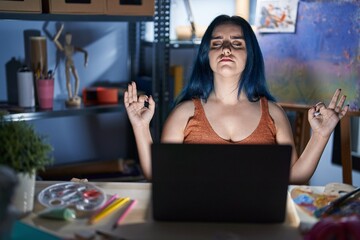 Young modern girl with blue hair sitting at art studio with laptop at night relax and smiling with eyes closed doing meditation gesture with fingers. yoga concept.