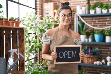 Hispanic woman working at florist holding open sign smiling looking to the side and staring away thinking.