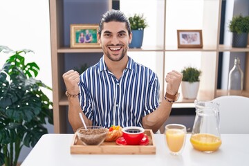 Hispanic man with long hair sitting on the table having breakfast screaming proud, celebrating victory and success very excited with raised arms