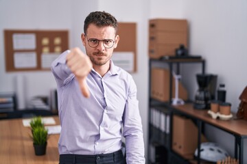 Young hispanic man at the office looking unhappy and angry showing rejection and negative with thumbs down gesture. bad expression.