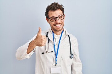 Young hispanic man wearing doctor uniform and stethoscope doing happy thumbs up gesture with hand. approving expression looking at the camera showing success.