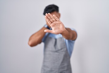 Fototapeta premium Hispanic young man wearing apron over white background covering eyes with hands and doing stop gesture with sad and fear expression. embarrassed and negative concept.