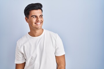 Hispanic man standing over blue background looking away to side with smile on face, natural expression. laughing confident.