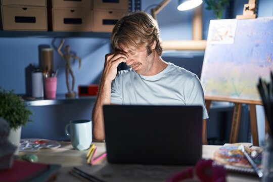 Middle age man sitting at art studio with laptop at night tired rubbing nose and eyes feeling fatigue and headache. stress and frustration concept. - Powered by Adobe