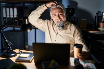 Middle age man with grey hair working at the office at night smiling confident touching hair with hand up gesture, posing attractive and fashionable