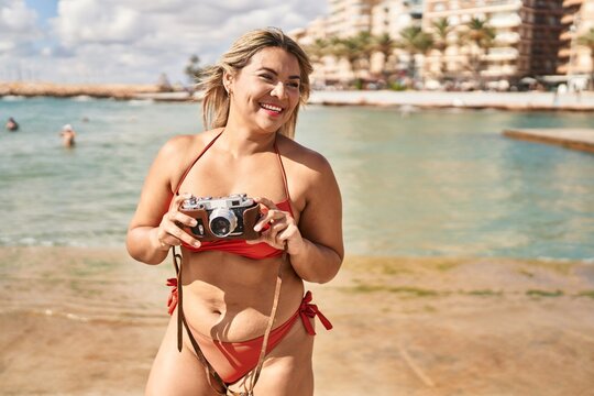 Young hispanic woman wearing bikini using vintage camera at seaside