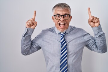 Hispanic business man with grey hair wearing glasses smiling amazed and surprised and pointing up with fingers and raised arms.