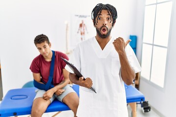 Young hispanic man working at pain recovery clinic with a man with broken arm surprised pointing with hand finger to the side, open mouth amazed expression.