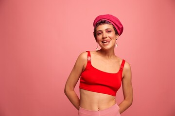 Obraz na płótnie Canvas Young athletic woman with a short haircut and purple hair in a red top and a pink hat with an athletic figure smiles and grimaces looking at the camera on a pink background