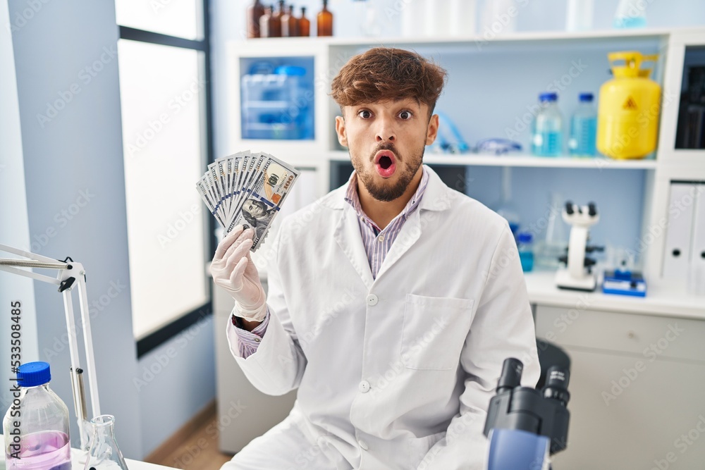 Sticker arab man with beard working at scientist laboratory holding money scared and amazed with open mouth 