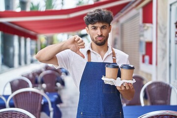 Arab man with beard wearing waiter apron at restaurant terrace with angry face, negative sign showing dislike with thumbs down, rejection concept