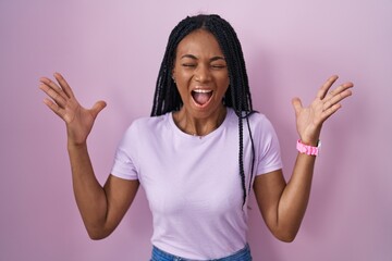 African american woman with braids standing over pink background celebrating mad and crazy for success with arms raised and closed eyes screaming excited. winner concept