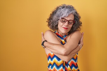 Middle age woman with grey hair standing over yellow background hugging oneself happy and positive,...