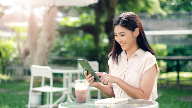 Asian business woman working outside freelancer using digital tablet device wireless remote communication technology, connectivity online worker casually enjoying outdoor in nature at cafe sunlight.