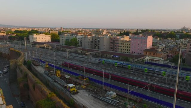 Slide and pan footage of modern high speed train and commuter unit leaving station on parallel tracks. Buildings in city at golden hour. Rome, Italy