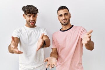 Young gay couple standing over isolated background smiling cheerful offering hands giving assistance and acceptance.