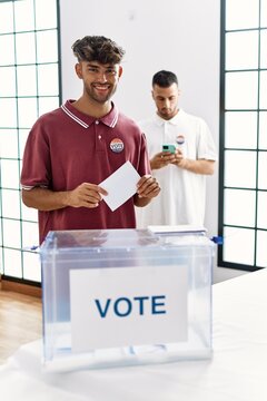 Young hispanic voter man smiling happy putting vote in ballot box at electoral center