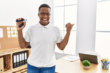 Young african man training muscle with hand grip at office pointing thumb up to the side smiling happy with open mouth