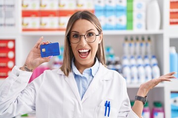 Young woman working at pharmacy drugstore holding credit card celebrating achievement with happy smile and winner expression with raised hand