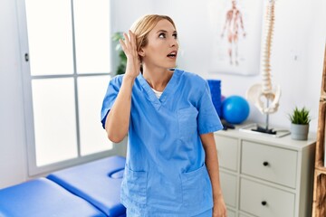 Beautiful blonde physiotherapist woman working at pain recovery clinic smiling with hand over ear listening an hearing to rumor or gossip. deafness concept.