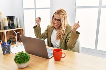 Beautiful blonde woman working at the office with laptop very happy and excited doing winner gesture with arms raised, smiling and screaming for success. celebration concept.