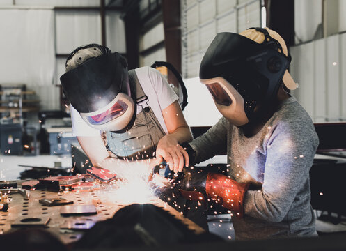 Industrial woman welder teaches younger student how to MIG weld metal with a torch