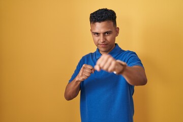 Young hispanic man standing over yellow background punching fist to fight, aggressive and angry...