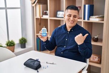 Hispanic young man holding glucometer device pointing thumb up to the side smiling happy with open mouth