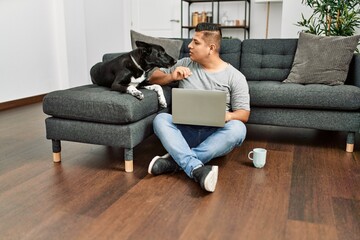Young hispanic man using laptop sitting on the floor with dog at home.