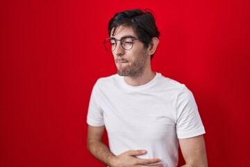 Young hispanic man standing over red background with hand on stomach because indigestion, painful illness feeling unwell. ache concept.
