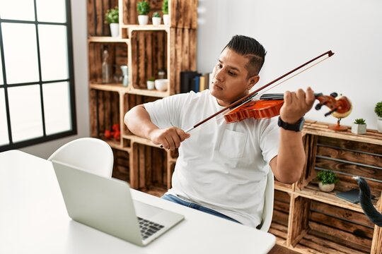 Young hispanic man having online violin lesson using laptop sitting on the table at home.