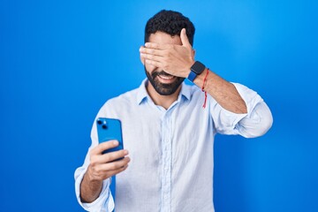 Hispanic man with beard using smartphone typing message smiling and laughing with hand on face...