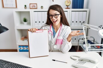 Obraz na płótnie Canvas Young doctor woman holding clipboard at the clinic celebrating achievement with happy smile and winner expression with raised hand
