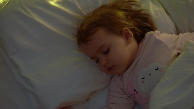 Cute Chuby Little Toddler Baby Girl 1-2 Years Old Sleeping In Pink Pajama On White Bed In Dark Bedroom. Child Have Relax Rest Time At Night Garland on Pillow. Childhood, Christmas, Parenthhood Concept