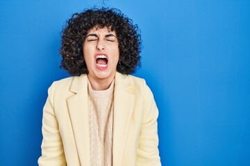 Fototapeta na wymiar Young brunette woman with curly hair standing over blue background angry and mad screaming frustrated and furious, shouting with anger. rage and aggressive concept.