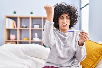 Young brunette woman with curly hair holding pregnancy test result annoyed and frustrated shouting with anger, yelling crazy with anger and hand raised