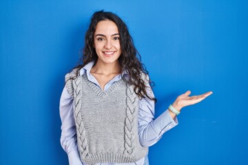 Young brunette woman standing over blue background smiling cheerful presenting and pointing with palm of hand looking at the camera.