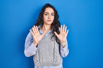Young brunette woman standing over blue background moving away hands palms showing refusal and...