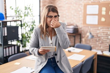 Young hispanic woman working at the office wearing glasses smelling something stinky and disgusting, intolerable smell, holding breath with fingers on nose. bad smell - obrazy, fototapety, plakaty