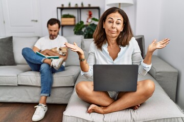 Hispanic middle age couple at home, woman using laptop clueless and confused expression with arms and hands raised. doubt concept.
