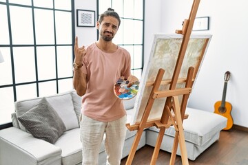 Young hispanic man with beard painting on canvas at home pointing up looking sad and upset,...