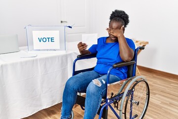 Young african woman sitting on wheelchair voting putting envelop in ballot box covering eyes with hand, looking serious and sad. sightless, hiding and rejection concept