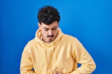 Fototapeta premium Hispanic man standing over blue background with hand on stomach because indigestion, painful illness feeling unwell. ache concept.
