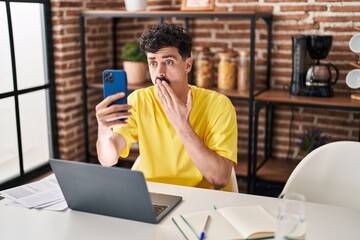 Hispanic man doing video call with smartphone covering mouth with hand, shocked and afraid for mistake. surprised expression