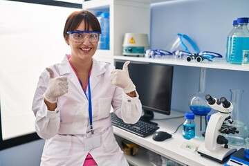 Young brunette woman working at scientist laboratory success sign doing positive gesture with hand, thumbs up smiling and happy. cheerful expression and winner gesture.