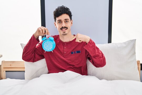 Hispanic man holding alarm clock in the bed pointing finger to one self smiling happy and proud