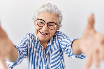 Senior woman with grey hair standing over white background looking at the camera smiling with open arms for hug. cheerful expression embracing happiness.