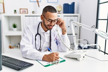 Fototapeta na wymiar Young hispanic man wearing doctor uniform speaking on the phone at clinic