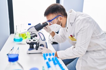 Young hispanic man scientist using microscope wearing medical mask at laboratory