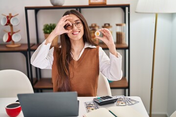 Young brunette woman working with laptop holding virtual currency bitcoin smiling happy doing ok...